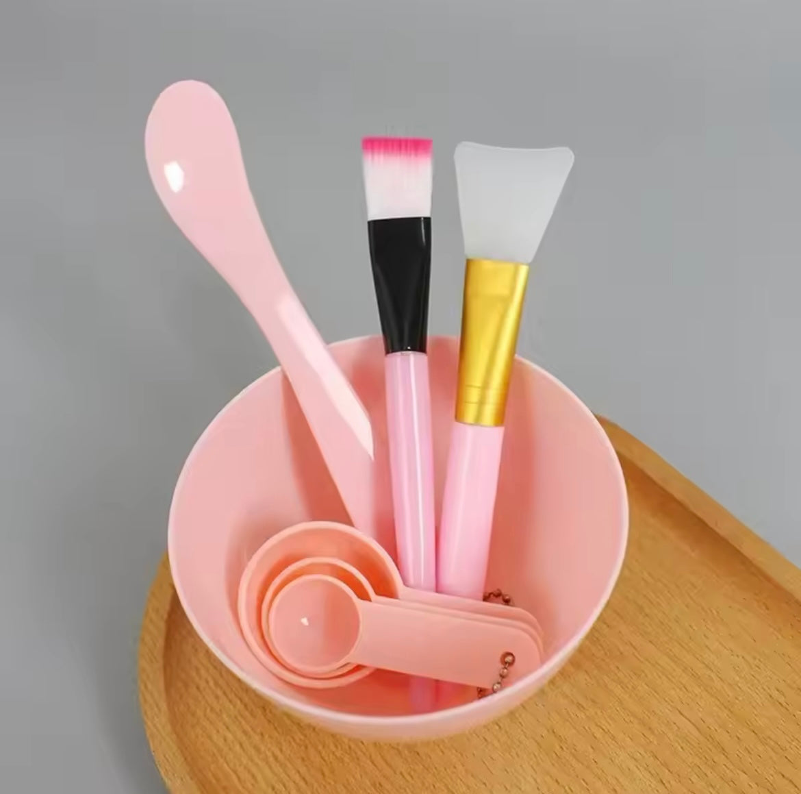 Set of cosmetic DIY face mask tools including a pink spoon, brushes, and measuring cups in a pink container on a wooden surface.