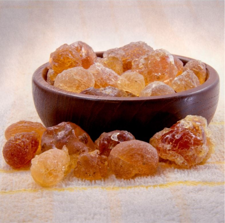 A bowl filled with golden-brown gum arabic beads on a light surface, with some beads scattered outside the bowl.